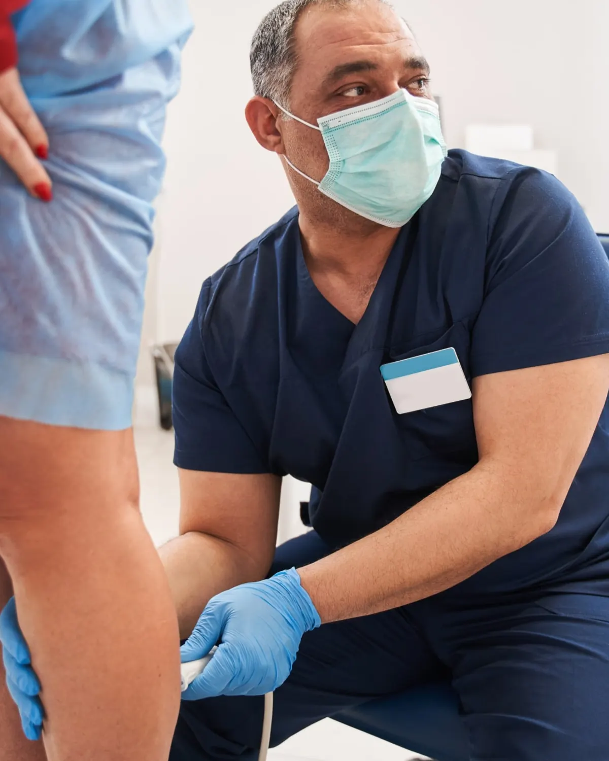 Masked clinician examining a patient's knee during treatment